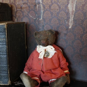 May include: A vintage teddy bear wearing a red outfit with white trim and a gold button. The bear is sitting on a black surface in front of a brown patterned wall.