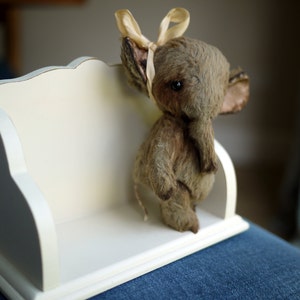 May include: A small, grey stuffed elephant with a cream-coloured bow sits on a white shelf with a decorative edge.