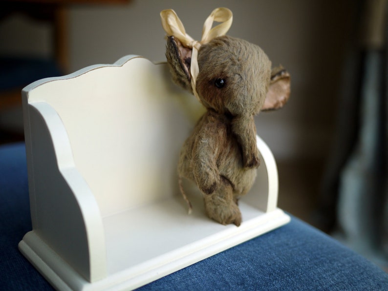 May include: A small, grey stuffed elephant with a cream-coloured bow sits on a white shelf with a decorative edge.