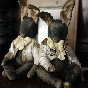 Könnte beinhalten: Zwei antike Plüschkaninchen-Puppen mit langen Ohren, gekleidet in cremefarbenen Jacken und Fliegen. Die Kaninchen haben dunkles Fell und sitzen, was eine Vintage-Ästhetik zeigt. Die Jacken haben goldene Knöpfe und einen kleinen Anhänger.