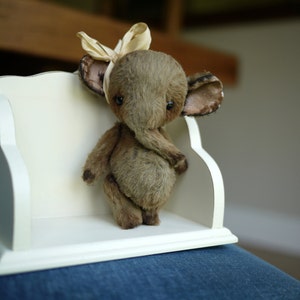 May include: A small, brown, plush elephant toy with a beige bow on its head. The elephant is sitting on a white shelf with a decorative edge.