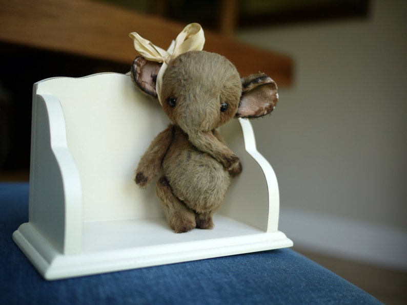 May include: A small, brown, plush elephant toy with a beige bow on its head. The elephant is sitting on a white shelf with a decorative edge.