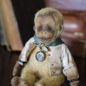 May include: A plush monkey toy wearing a white shirt with a blue collar and a gold chain with a small photo charm. The monkey has a brown face and brown fur.