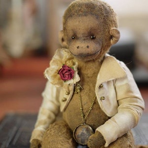 Może przedstawiać: Pluszowa zabawka małpy w stylu vintage, ubrana w białą kurtkę ze złotymi guzikami i złoty łańcuszek z zawieszką. Małpa ma brązowe futro i czerwoną różę na kurtce.