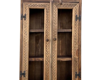 May include: A wooden cabinet with two glass doors. The cabinet is made of dark brown wood with intricate carvings around the edges and a decorative starburst design at the top. The doors have small, ornate hinges.