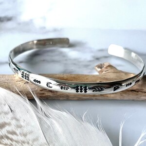 May include: A silver bangle bracelet with a hammered finish and an engraved design of arrows, moons, and dots.