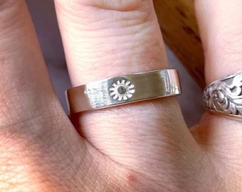 May include: Two silver rings on a finger. The ring on the index finger has a floral design and a round center with an inscription. The ring on the middle finger has a simple band with a small circular design.