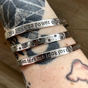 May include: Three silver bracelets stacked on a person's arm. The top bracelet has the text "you have no power over me". The middle bracelet has a pattern of stars. The bottom bracelet has the text "when it rains look for rainbows".