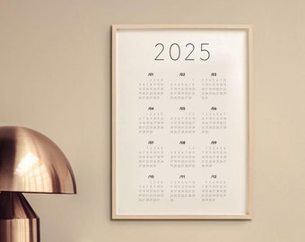 Puede incluir: Un calendario enmarcado para el año 2025 con cada mes mostrado en un formato de cuadrícula. El calendario está colgado en una pared beige sobre una cómoda de madera con una lámpara dorada, una escultura de coral blanco y un libro.