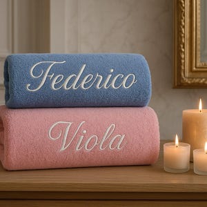 Op de afbeelding: Twee opgerolde handdoeken, één blauw en één roze, gestapeld op een houten oppervlak. De blauwe handdoek heeft de naam "Federico" in het wit geborduurd, en de roze "Viola". Brandende kaarsen op de achtergrond.