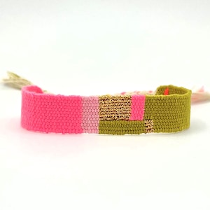 May include: A woven friendship bracelet with a vibrant color scheme. The bracelet features sections of bright pink, light pink, gold, and olive green. The bracelet is approximately 1 inch wide.