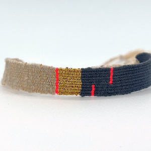 Könnte beinhalten: Ein gewebtes Armband mit beige, goldenen und schwarzen Bändern. Das Armband hat einen roten Faden, der in das schwarze Band eingewebt ist.