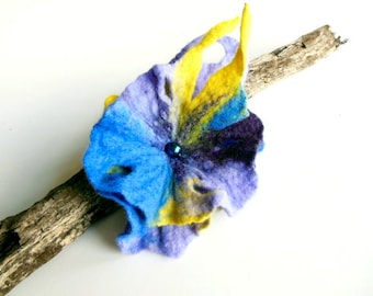 May include: A blue, yellow, and purple felt flower with a blue bead in the center. The flower is sitting on a piece of driftwood.