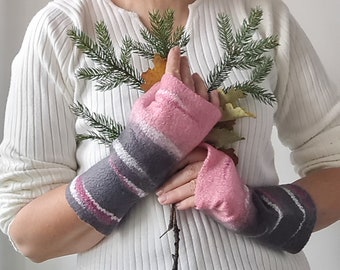 Può includere: Un paio di guanti senza dita con un aspetto in lana infeltrita e testurizzata, caratterizzati da sezioni orizzontali a blocchi di colore in rosa tenue, grigio scuro e sottili strisce bianche. I guanti coprono dalle nocche all'avambraccio.