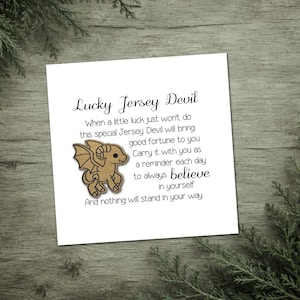 May include: A white card with the text "Lucky Jersey Devil" and a cartoon illustration of a Jersey Devil. The text includes the phrases "good fortune" and "believe in yourself". The card is on a wooden surface.