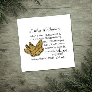 May include: A white card with the text "Lucky Mothman" and a gold-colored moth illustration. The card includes text about good fortune and believing in yourself. The card is on a wooden surface with greenery.