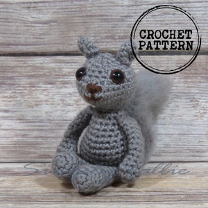 May include: A gray crocheted squirrel amigurumi toy with brown eyes and a brown nose. The toy is sitting on a wooden surface. CROCHET PATTERN is written in a circle on the image.