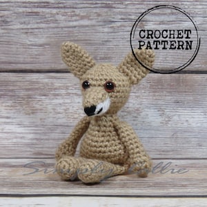 May include: Crochet pattern for a small, tan-colored fox amigurumi. The fox is sitting with its ears perked up and has brown eyes and a black nose.