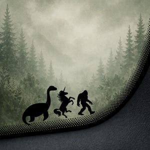May include: A black silhouette design featuring a Loch Ness Monster, a unicorn, and Bigfoot walking in front of a forest scene. The background shows a misty forest with tall evergreen trees. The design is set against a gray background.