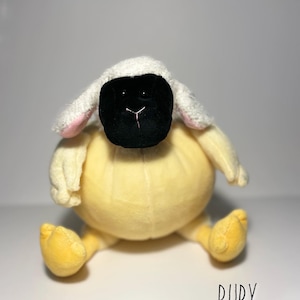 May include: A yellow plush sheep with a black face and white fur on its head. The sheep is sitting with its legs crossed and has the word "RUBY" written below it.