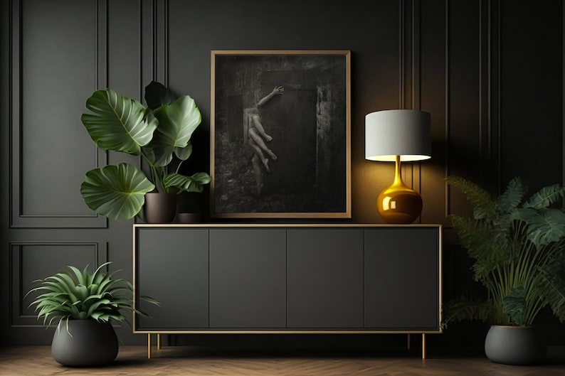 May include: A black cabinet with gold accents sits in a dark room with a framed black and white photograph of a woman reaching up towards a doorway.  The cabinet is flanked by two potted plants, one with large green leaves and the other with smaller, fern-like leaves. A gold table lamp with a white shade sits on the cabinet.