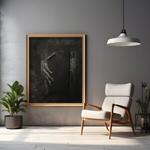 May include: A black and white photograph of a person reaching out from a dark, shadowy space. The photograph is framed in a wooden frame and hanging on a gray wall.