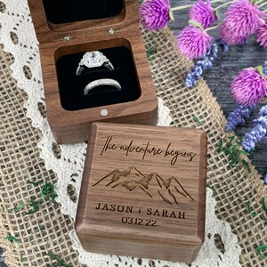 May include: A wooden ring box with a hinged lid. The box is engraved with the text "The adventure begins" and a mountain range. The box also has the names "Jason + Sarah" and the date "03.12.22" engraved on it.