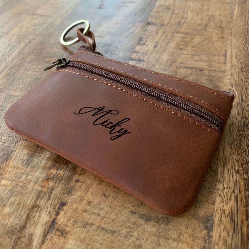 May include: A brown leather coin purse with a zipper closure and a key ring. The purse is personalized with the name "Micky" in cursive.