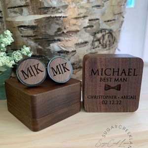 May include: A wooden box with a lid, engraved with the text "MICHAEL BEST MAN CHRISTOPHER + ABIGAIL 02.12.22". The box contains two wooden cufflinks engraved with the initials "MK".