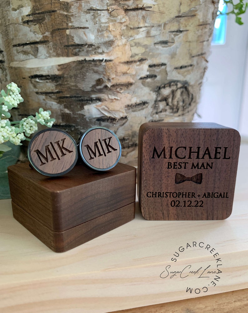 May include: A wooden box with a lid, engraved with the text "MICHAEL BEST MAN CHRISTOPHER + ABIGAIL 02.12.22". The box contains two wooden cufflinks engraved with the initials "MK".