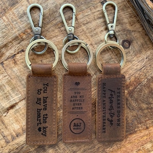 May include: Three brown leather keychains with metal rings and clasps. The keychains have engraved messages: "You have the key to my heart", "You are my happily ever after A&J 12.15.18", and "3 Years Down Forever to go. Happy Anniversary".