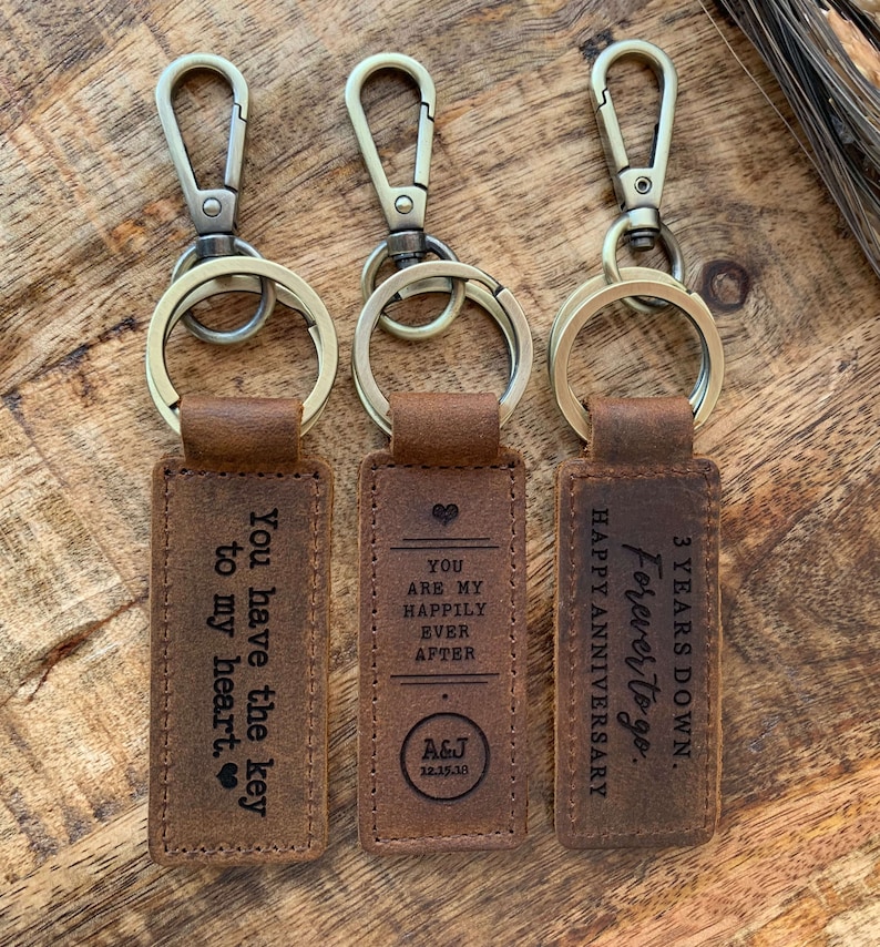 May include: Three brown leather keychains with metal rings and clasps. The keychains have engraved messages: "You have the key to my heart", "You are my happily ever after A&J 12.15.18", and "3 Years Down Forever to go. Happy Anniversary".