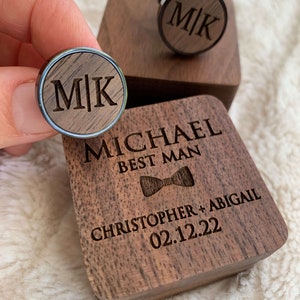 May include: A wooden box with a bowtie design and the text "MICHAEL BEST MAN" and "CHRISTOPHER + ABIGAIL 02.12.22". The box contains two black cufflinks with a wood inlay and the initials "M|K".