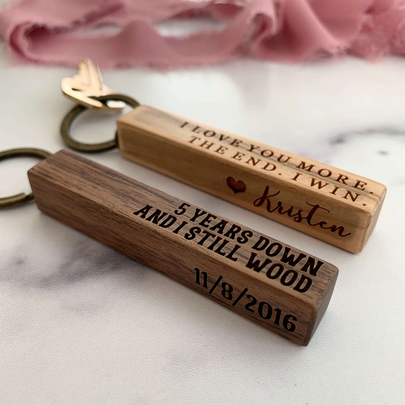 May include: Two wooden keychains with engraved messages. The first keychain is light brown and reads "I love you more. The end. I win. Kristen". The second keychain is dark brown and reads "5 years down and I still wood 11/8/2016".