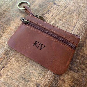 May include: A brown leather key chain with a zippered pouch and a key ring. The pouch has a monogram of the initials "K|V" on the front.