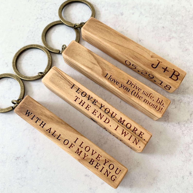 May include: Four wooden keychains with engraved messages. The keychains are made of light brown wood and have a brass key ring. The messages on the keychains are: "I love you more. The end. I win.", "I love you with all of my being", "Drive safe, bb. I love you (the most)!", and "J+B 05.29.18".