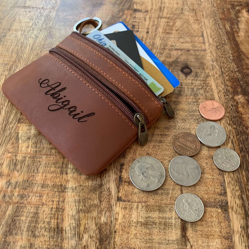 May include: A brown leather coin purse with a zipper closure and the name "Abigail" engraved on the front. The purse is open and contains a few credit cards and some US coins.