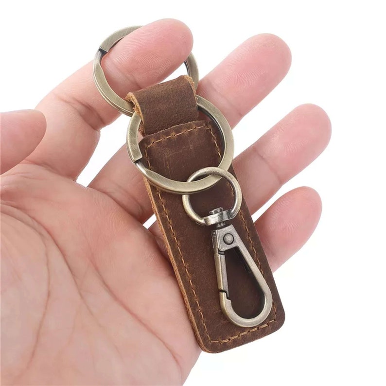 May include: A brown leather key chain with a metal ring and a metal clip. The key chain is in a hand.