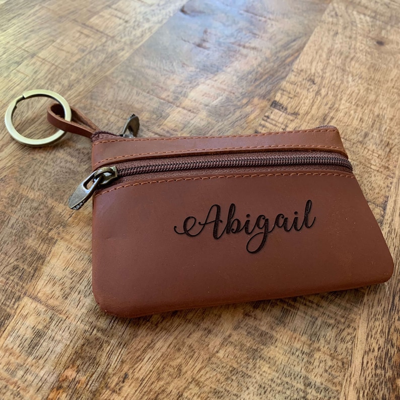 May include: A brown leather coin purse with a zipper closure and a key ring. The purse is personalized with the name "Abigail" in a cursive font.