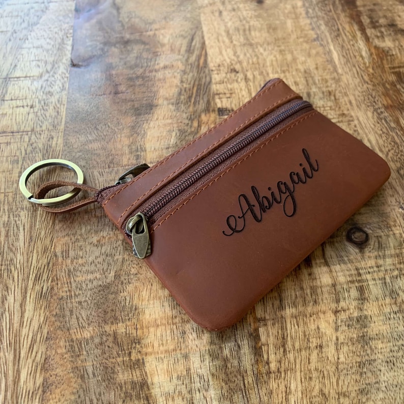 May include: Brown leather coin purse with a zipper closure and a key ring. The purse is personalized with the name "Abigail" in a cursive font.