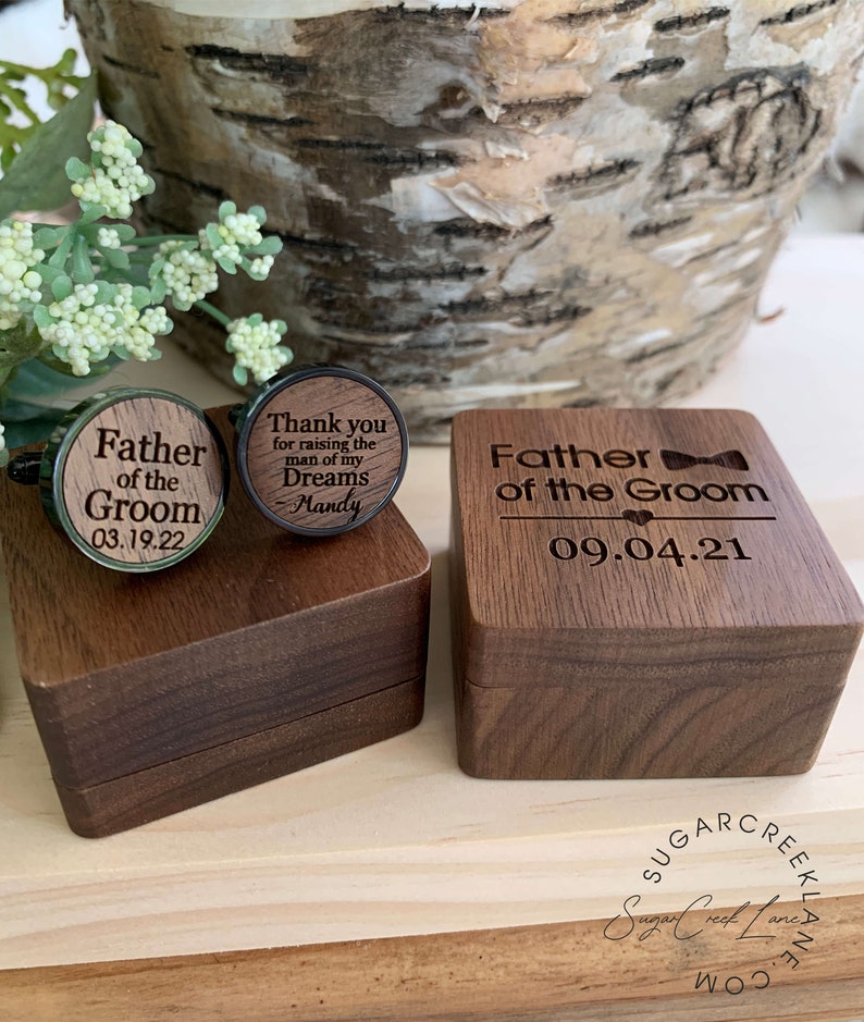 May include: Wooden box with a bow tie design and the text "Father of the Groom" and the date "09.04.21". The box is open and contains two wooden cufflinks. One cufflink has the text "Father of the Groom" and the date "03.19.22". The other cufflink has the text "Thank you for raising the man of my Dreams Mandy".