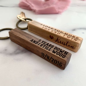 May include: Two wooden keychains with engraved messages. The first keychain is light brown and reads "I love you more the end. I win. Kristen". The second keychain is dark brown and reads "5 years down and I still wood 11/8/2016".