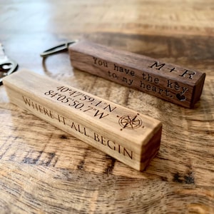 May include: Two wooden keychains with engraved text. The first keychain is brown and reads "M+R You have the key to my heart." The second keychain is light brown and reads "40°47'59.1"N 84°05'50.7"W WHERE IT ALL BEGIN".