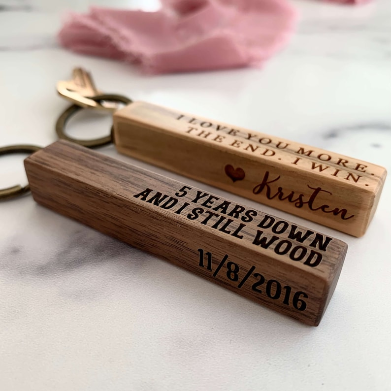 May include: Two wooden keychains with engraved messages. The first keychain is light brown and reads "I love you more. The end. I win. Kristen". The second keychain is dark brown and reads "5 years down and I still wood 11/8/2016".