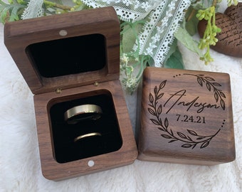 May include: A wooden ring box with a black velvet interior. The box is open and two gold wedding bands are inside. The lid of the box is closed and has the text "Anderson 7.24.21" engraved on it.