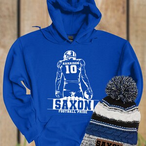 May include: Royal blue hoodie with a white silhouette of a football player wearing jersey number 10 with the name "HARRISON" above the number. The hoodie has the words "SAXON FOOTBALL PRIDE" printed below the player. A blue, white, and black striped beanie with a white pom pom is next to the hoodie. The beanie has a brown patch with the words "SAXON FOOTBALL PRIDE" printed on it.