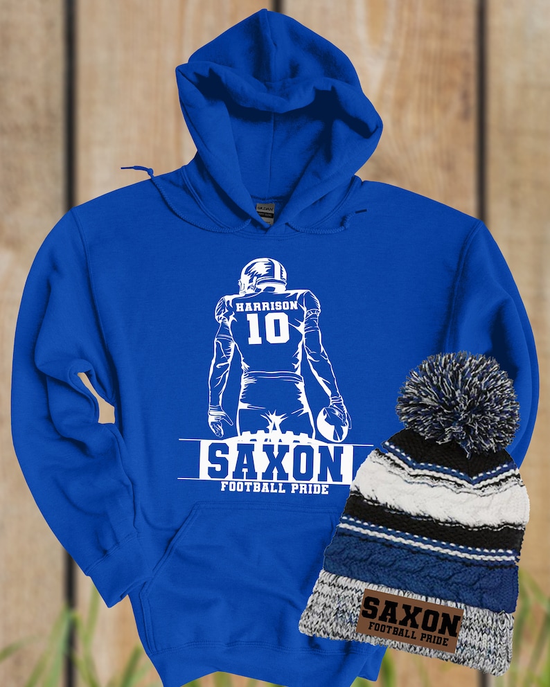 May include: Royal blue hoodie with a white silhouette of a football player wearing jersey number 10 with the name "HARRISON" above the number. The hoodie has the words "SAXON FOOTBALL PRIDE" printed below the player. A blue, white, and black striped beanie with a white pom pom is next to the hoodie. The beanie has a brown patch with the words "SAXON FOOTBALL PRIDE" printed on it.