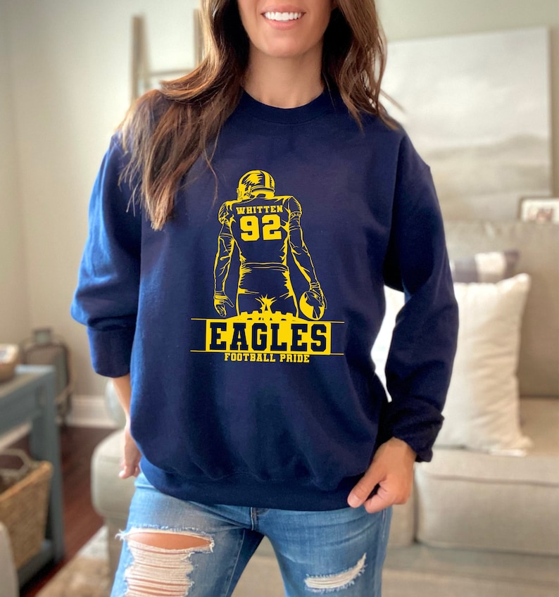 May include: A navy blue sweatshirt with a yellow graphic of a football player with the text "Whitten 92" on the back. The graphic also includes the text "Eagles Football Pride".