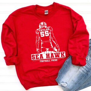 May include: Red sweatshirt with a white graphic of a football player with the number 55 and the text "TAYLOR" above the number. The text "SEA HAWK" is below the player with the text "FOOTBALL PRIDE" below that.