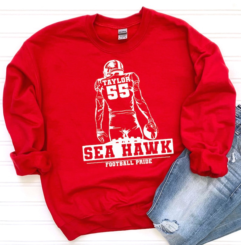 May include: Red sweatshirt with a white graphic of a football player with the number 55 and the text "TAYLOR" above the number. The text "SEA HAWK" is below the player with the text "FOOTBALL PRIDE" below that.